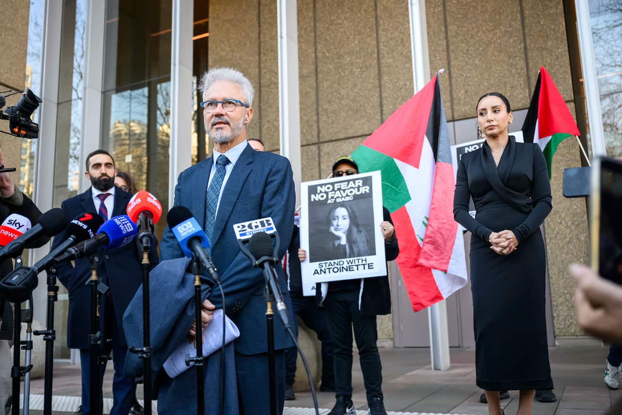 A man wearing a suit, tie and glasses, speaking to journalists in front of microphones.