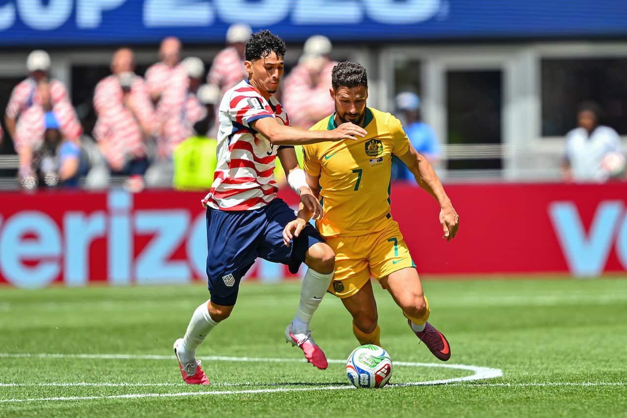 A full shot of two soccer players intensely competing for the ball on a green field during a match. The player on the left, representing the US, wears a white jersey with red horizontal stripes, dark blue shorts, and white socks. The player on the right, representing Australia, wears a solid yellow jersey with the number 7 and matching yellow shorts. Both players are closely matched, leaning into each other as they vie for possession of the white, green, and red soccer ball at their feet. The background features a brightly lit stadium with blurred spectators and a red perimeter advertisement banner.