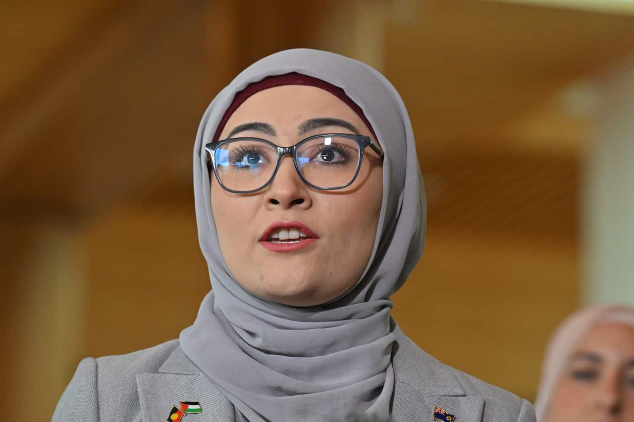 A woman in glasses and a grey hijab is speaking. 