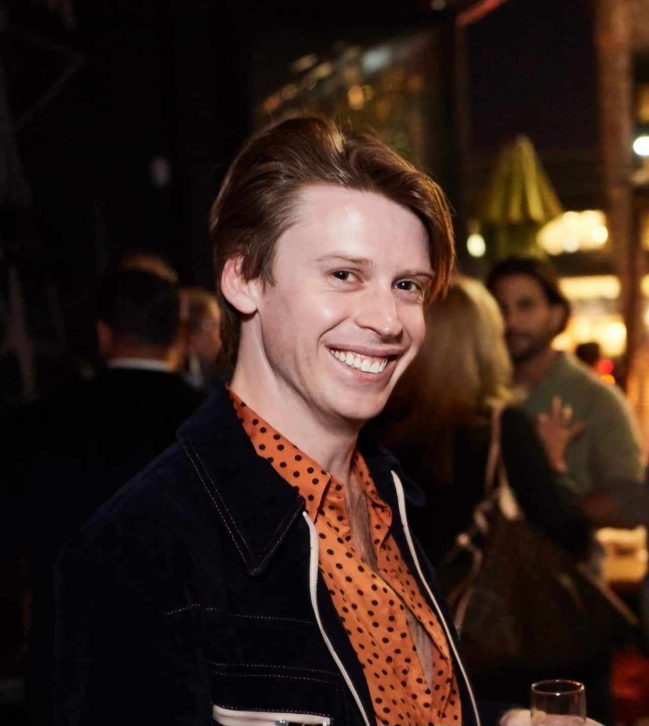 A man smiling wearing an orange shirt and a black jacket. 