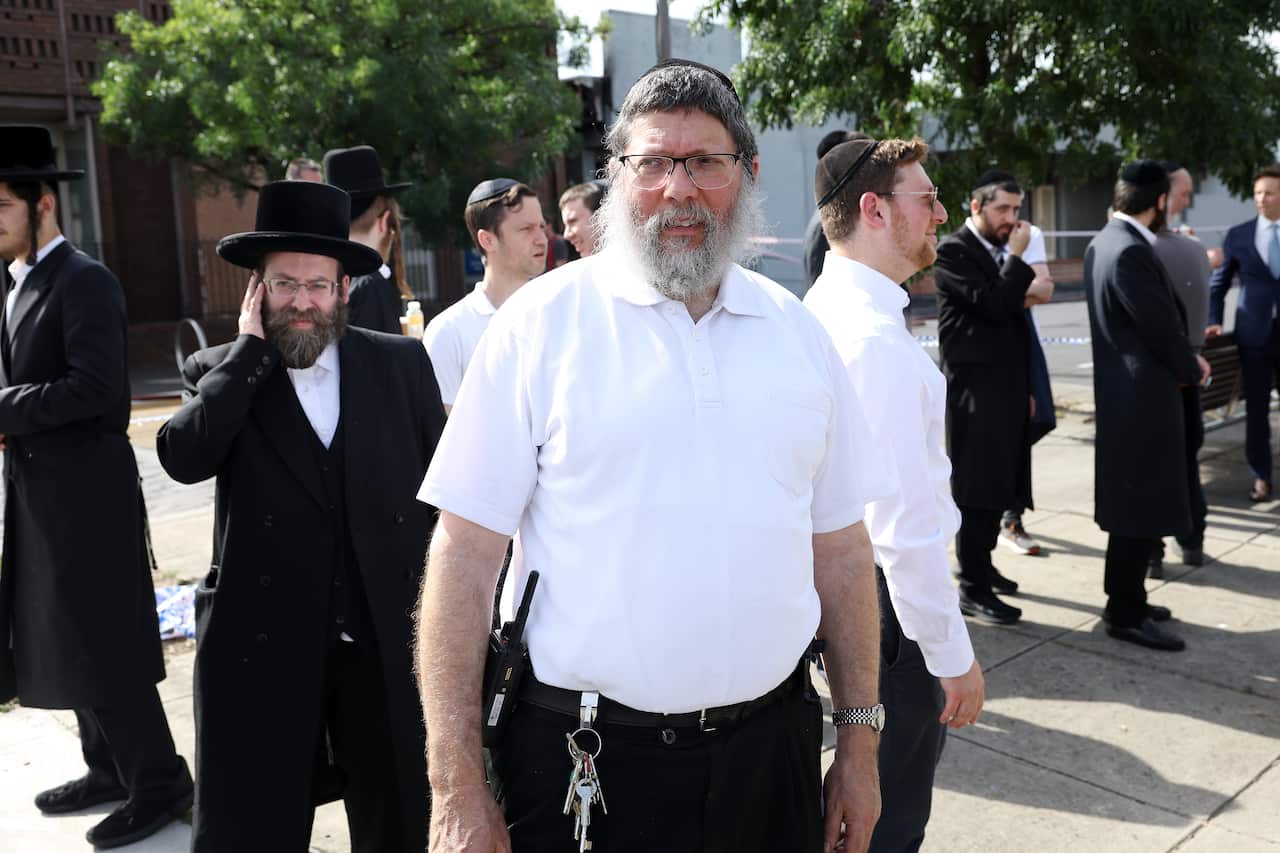 A man in a white shirt and kippah looks ahead while several men in long black coats and matching hats stand scattered in the background.