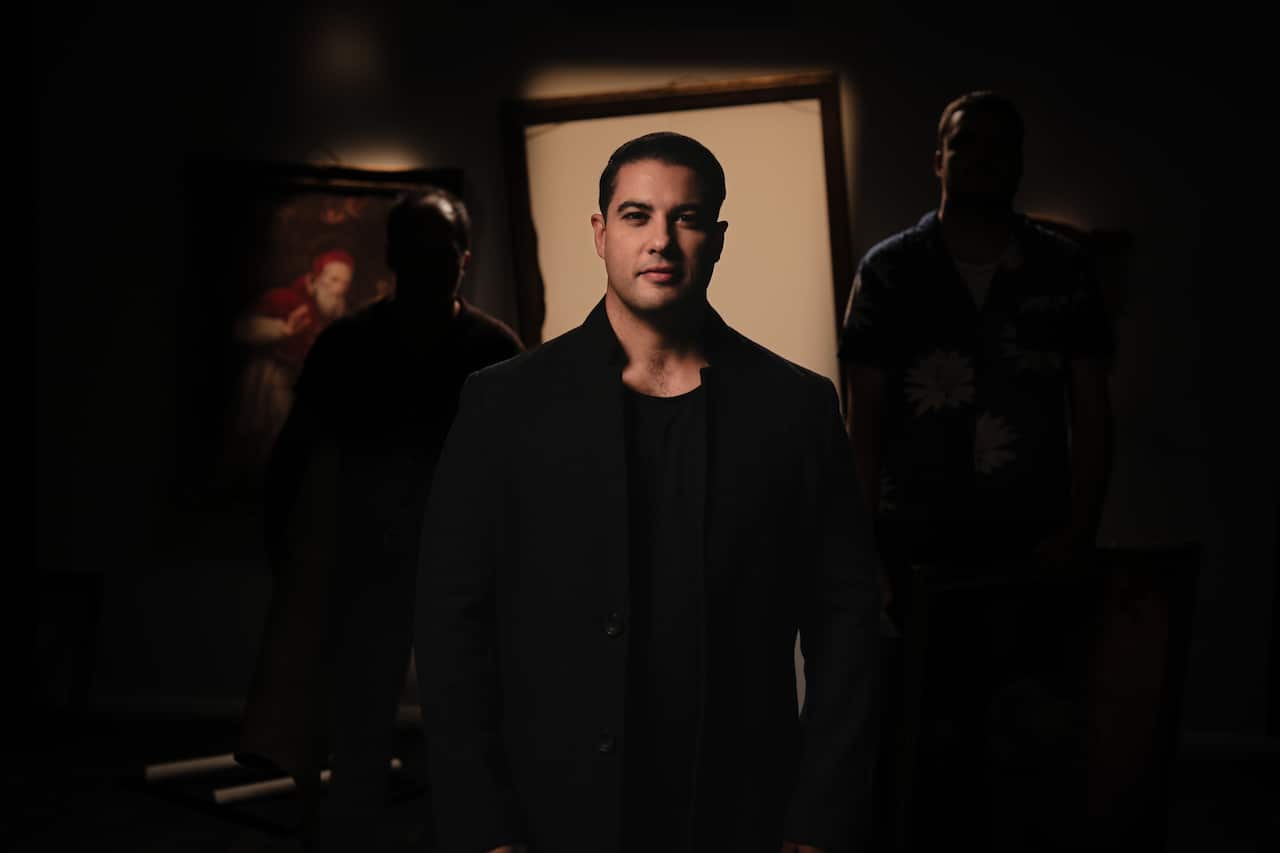 Marc Fennell stands in a gallery with 2 figures in silhouette behind him