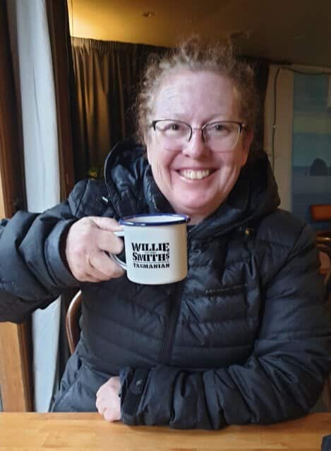 A smiling woman in a black puffer jacket sits at a table and holds an enamel mug featuring the "Willie Smiths Tasmanian" logo.