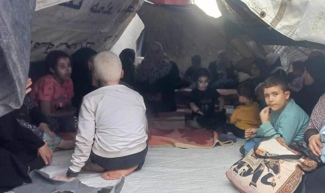 A group of women and children are gathered together, sitting on mats inside a large makeshift tent or tarp-covered shelter.