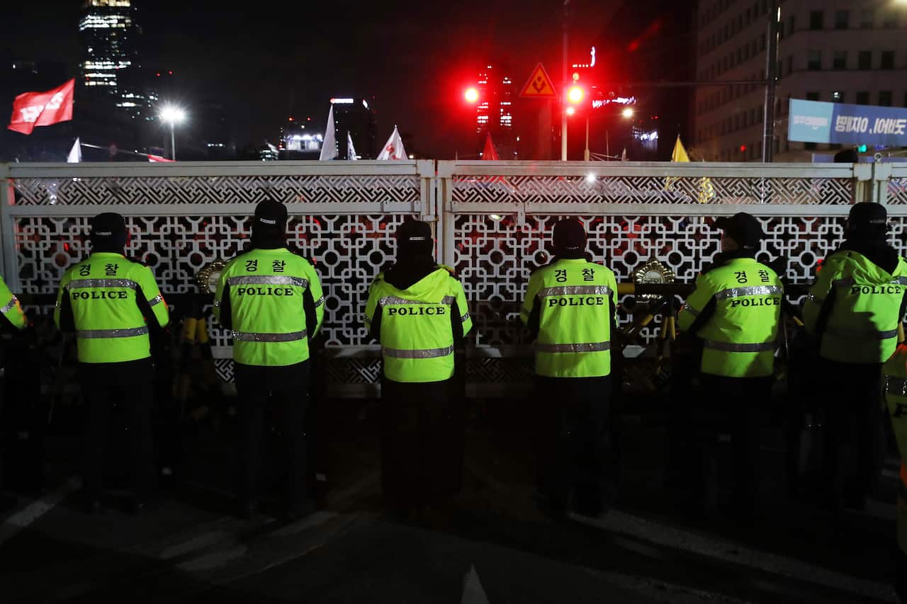 Police in hi vis vests stand in front of a gate at night.