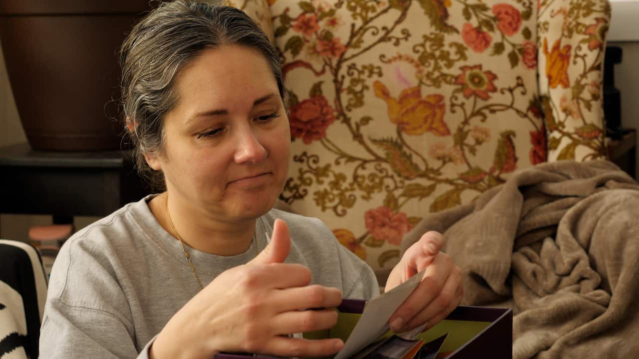 A woman looks through pictures in a box. Behind her is a floral armchair. 