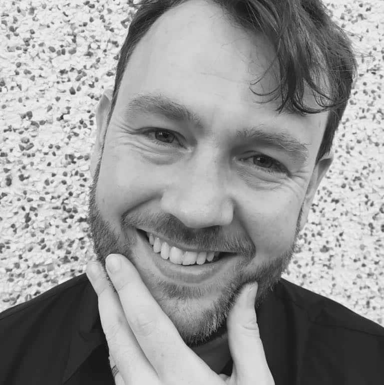 A black and white photo of a man in his 40s, smiling and touching his chin