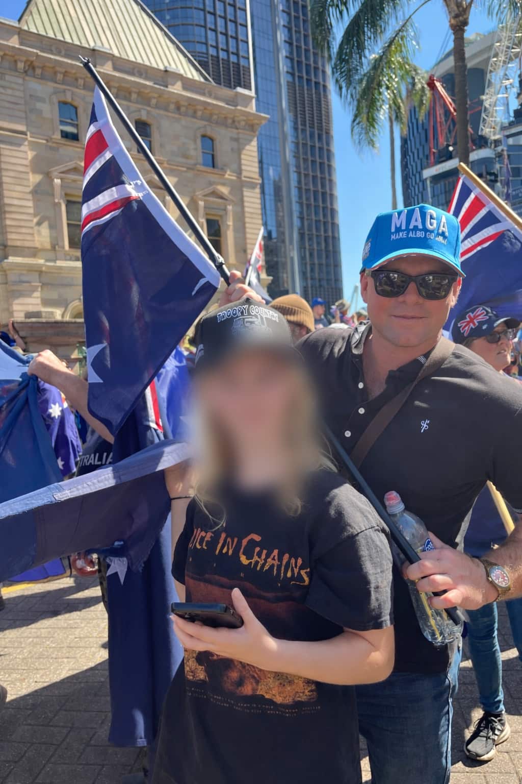 A man wearing a blue "MAGA" style hat and sunglasses stands next to a person in an Alice in Chains T-shirt at a sunny outdoor rally featuring Australian flags and historical stone architecture.