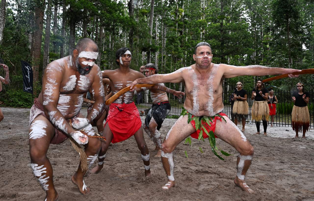Four male Aboriginal dancers with white paint markings on their bodies. There are four women dancers wearing grass skirts and black tops in the background  