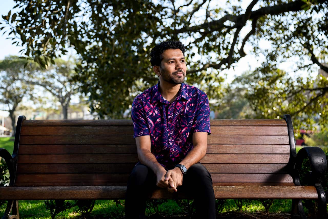 Vishal Prasad sits on a park bench, under a large tree, wearing a bright purple button-up shirt.