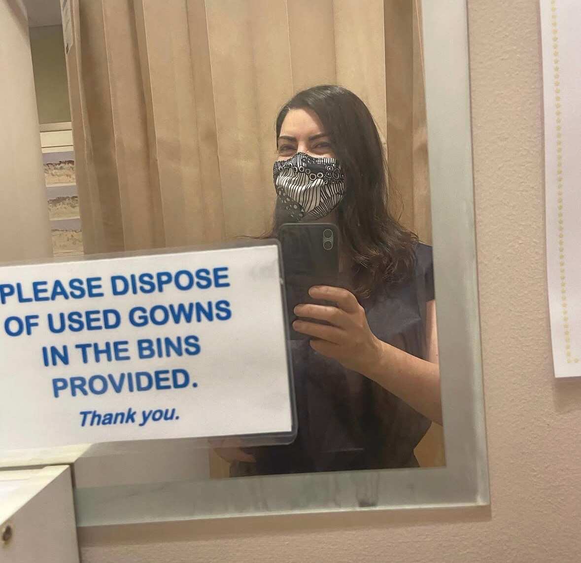 A woman taking a mirror selfie in a hospital bathroom. There's a sticker on the mirror with the words: "Please dispose of used gowns in the bins provided. Thank you."