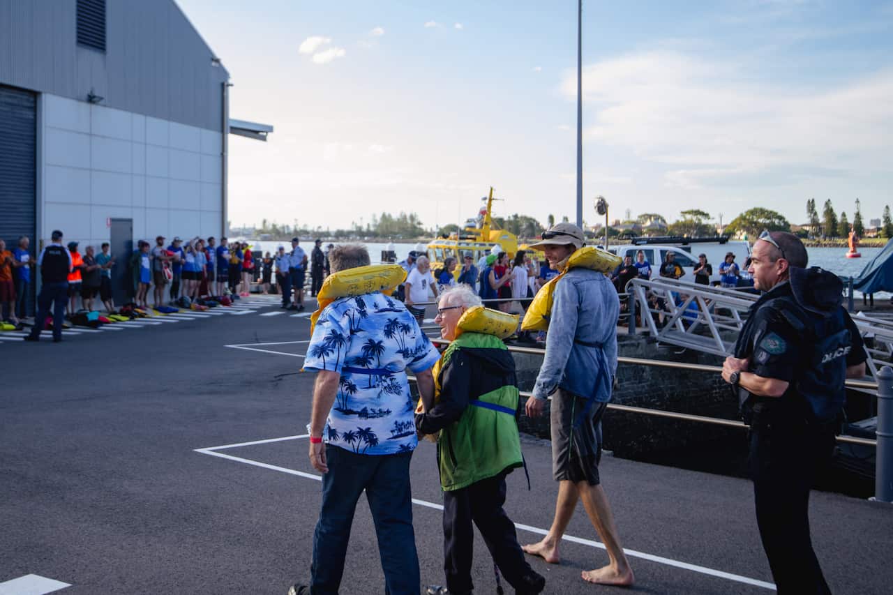 People wearing lifejackets on land next to the sea at a port.