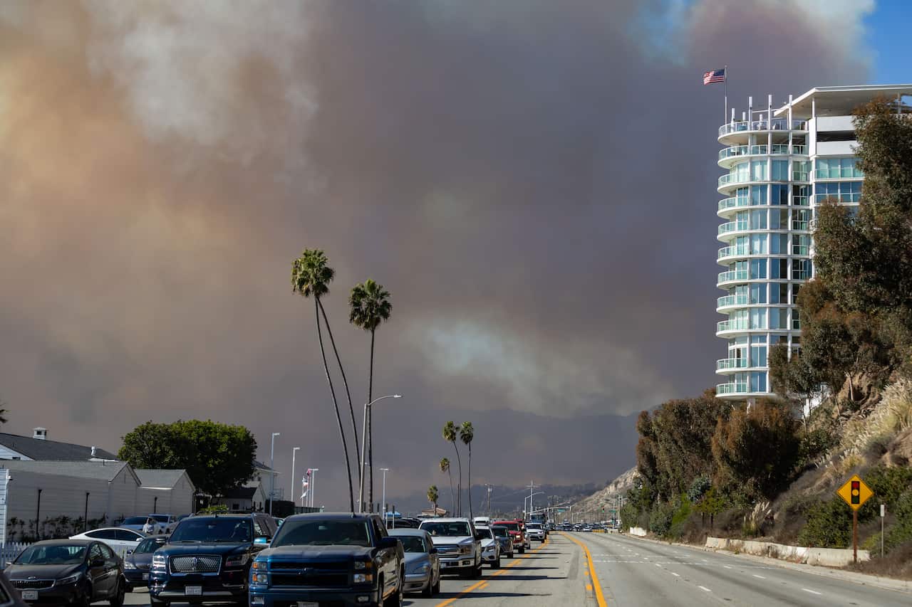 A line of cars on a street, with smoke rising from fires in the background.