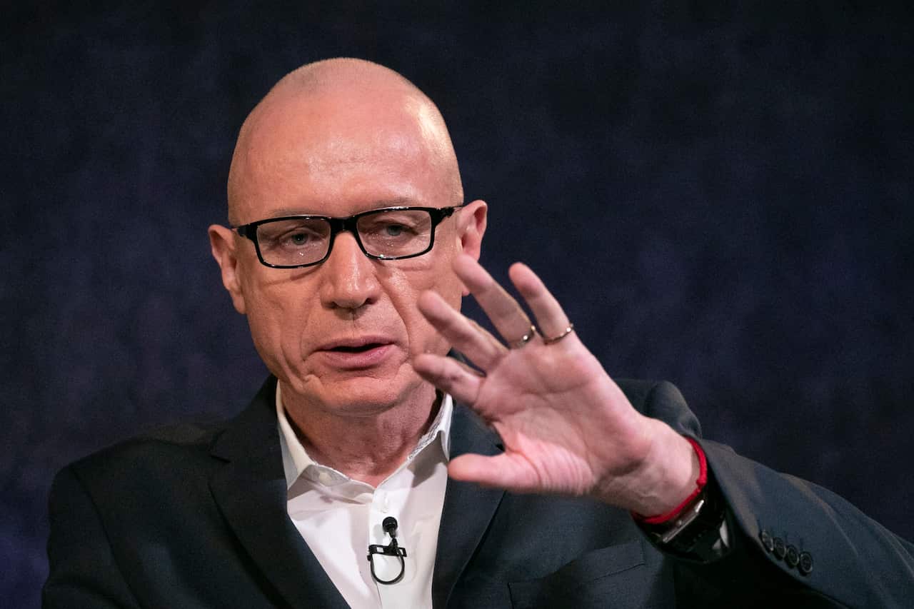A man wearing a suit and glasses gestures at a press conference. 