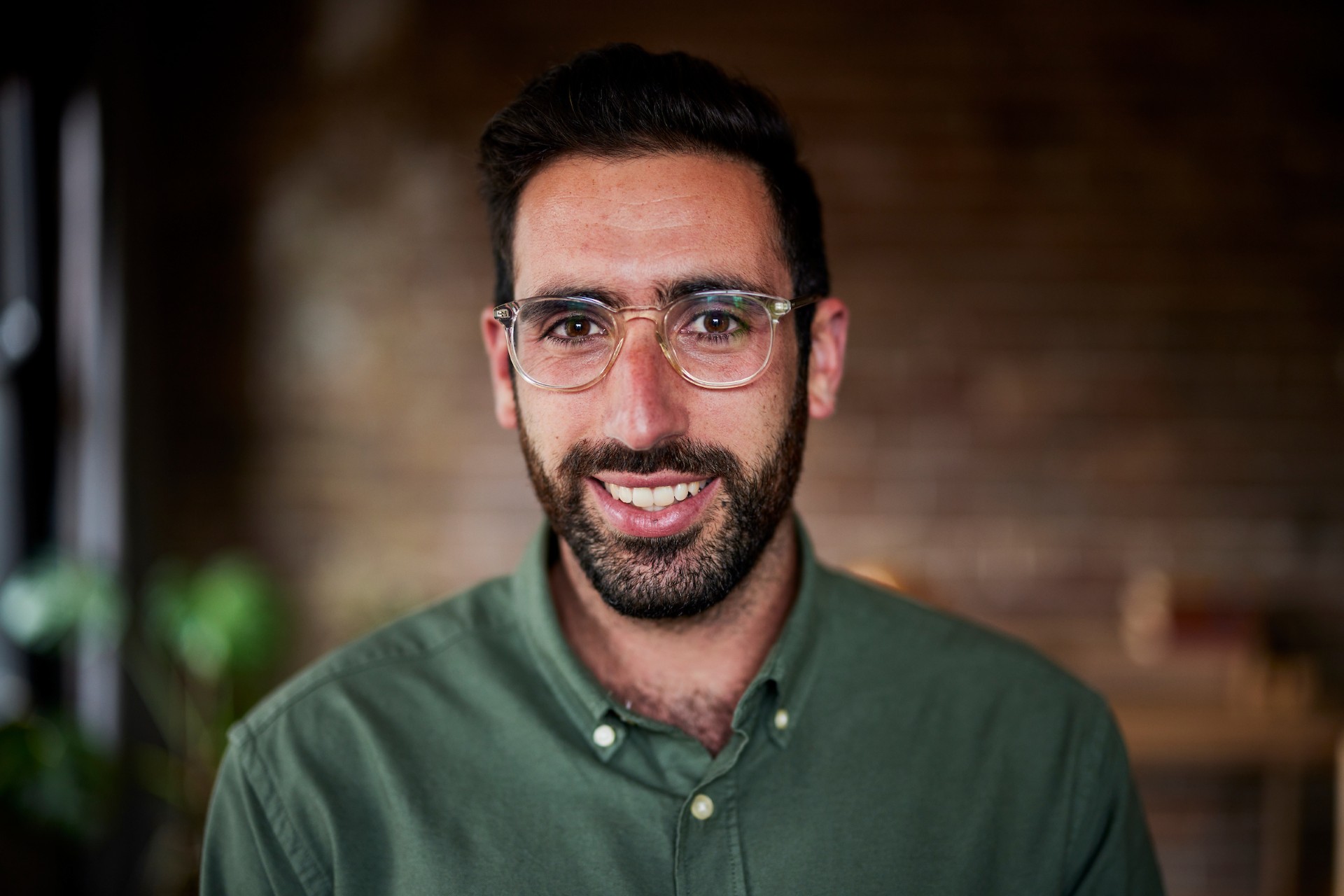A man wearing a green shirt is smiling. He has short dark hair, a beard and glasses.