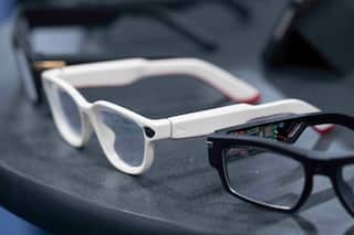 A pair of black and white glasses sit on a grey table. A small in built camera and wires can be seen on the glasses.