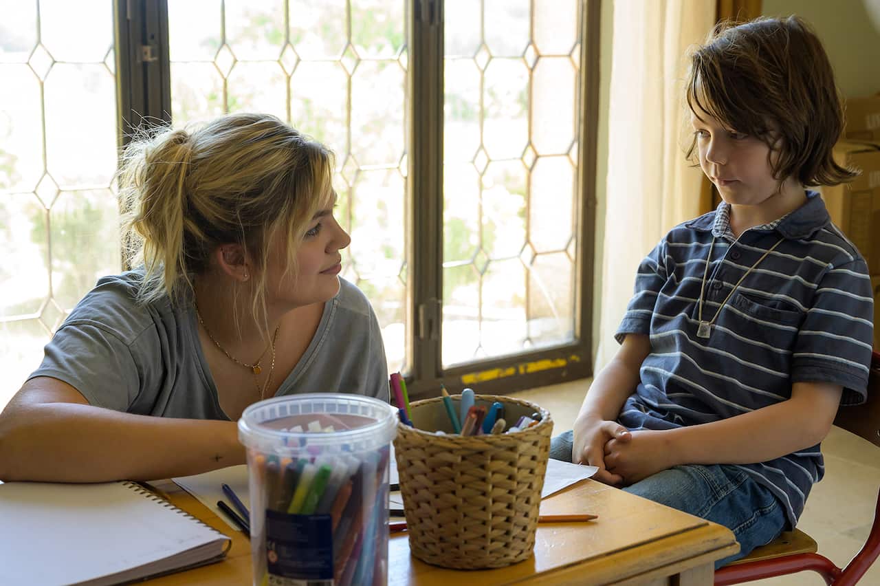 A child sits at a low table. A woman with blond hair is on the floor, leaning on the table, and looking at the child. Pencils and paper can be seen on the table. 