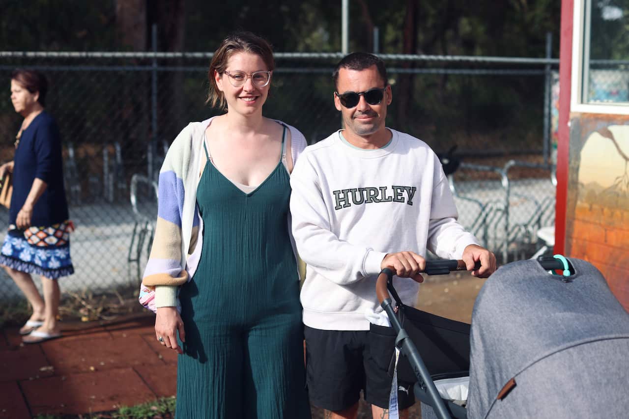 A young couple pushing a pram pose for a photo in front of a wire-mesh fence. 