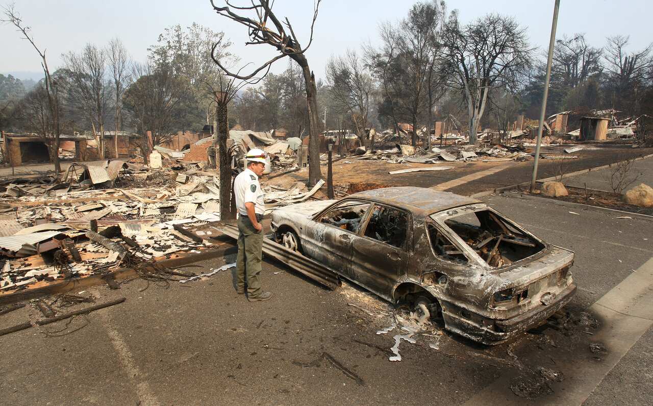 A man stands looking at a burned out car.