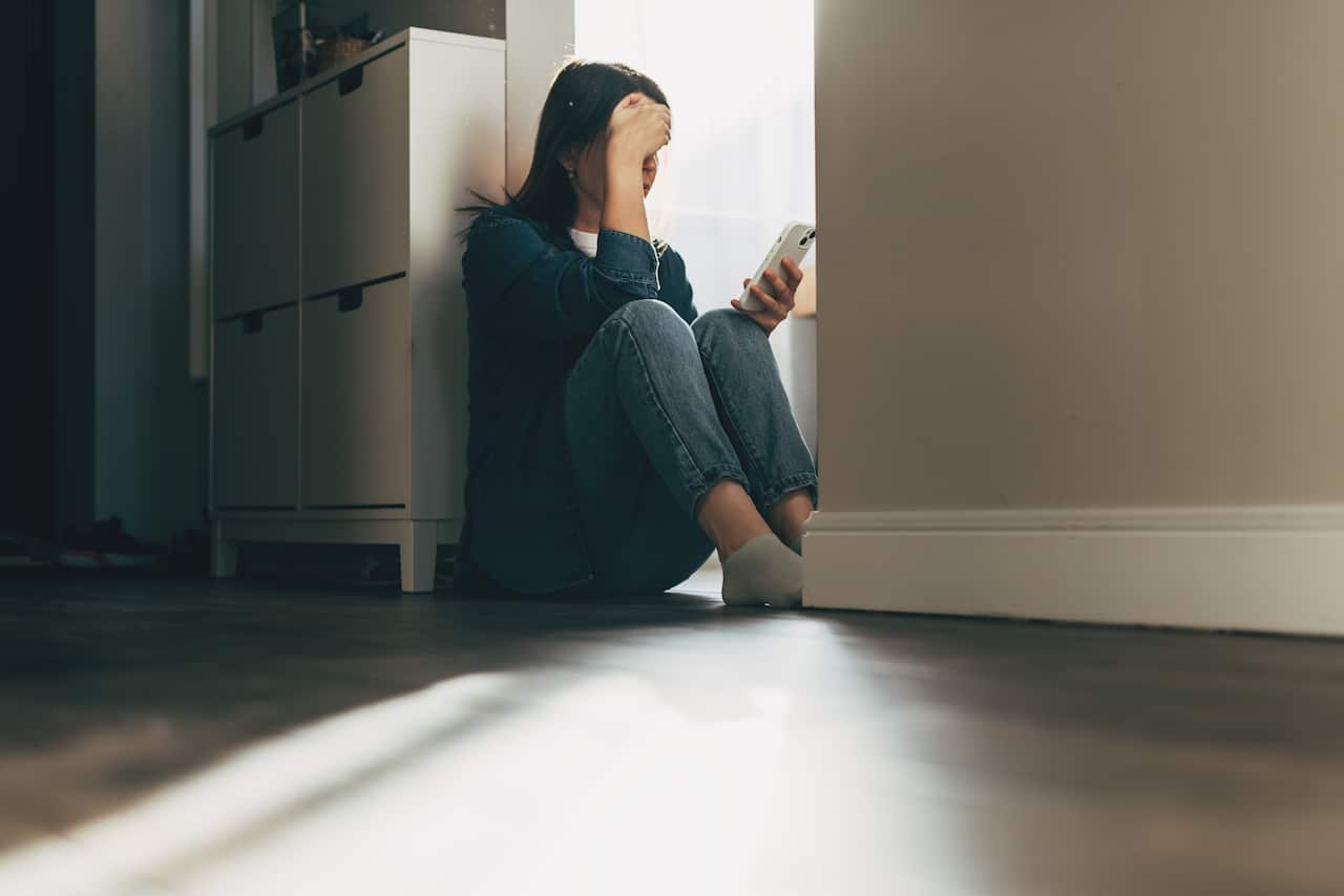 Silhouette of a woman sitting on the floor at home, holding a phone in one hand and resting her other hand on her forehead.