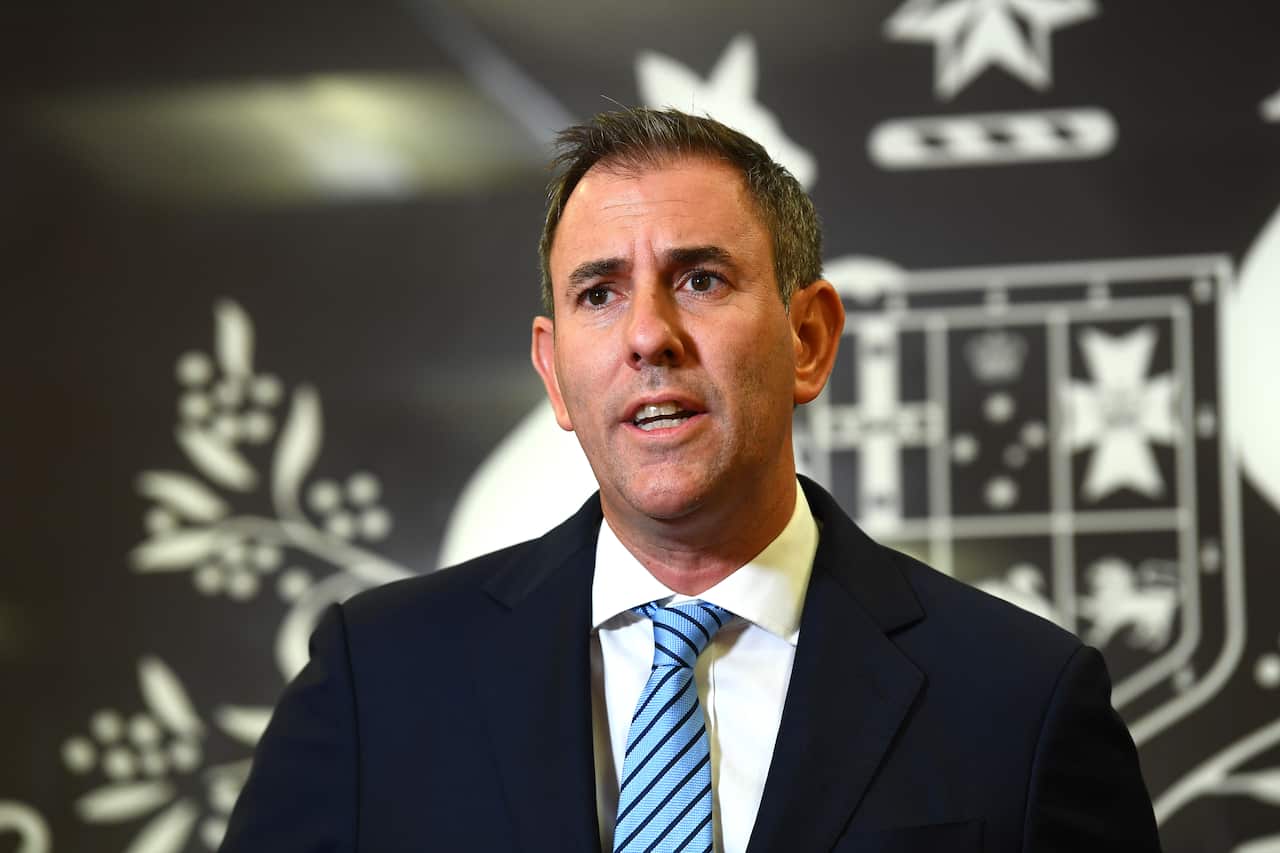 A man in a black suit standing in front of a coat of arms background.