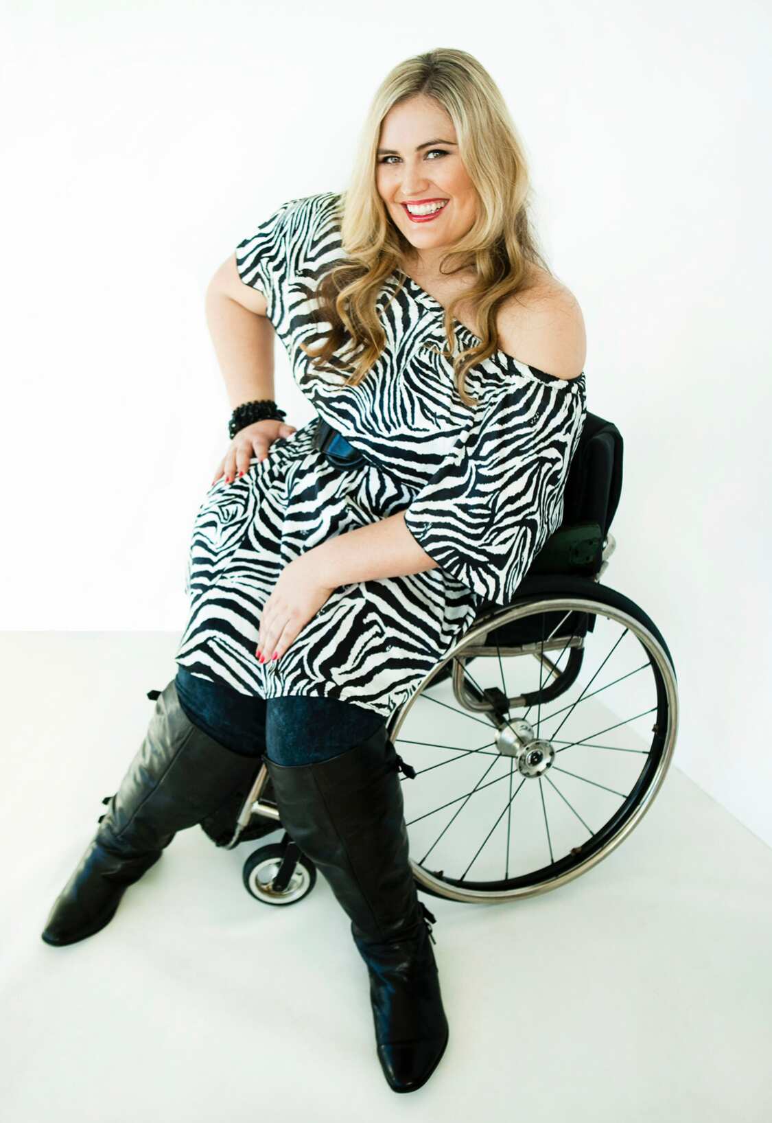 A woman wearing black knee high boots and a zebra print dress, seated in a wheelchair and smiling.