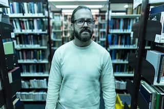 A man with glasses, a beard, and tied back hair and wearing a white long-sleeved t-shirt stands in the middle of an underground library. The light casts a blue glow over the scene.
