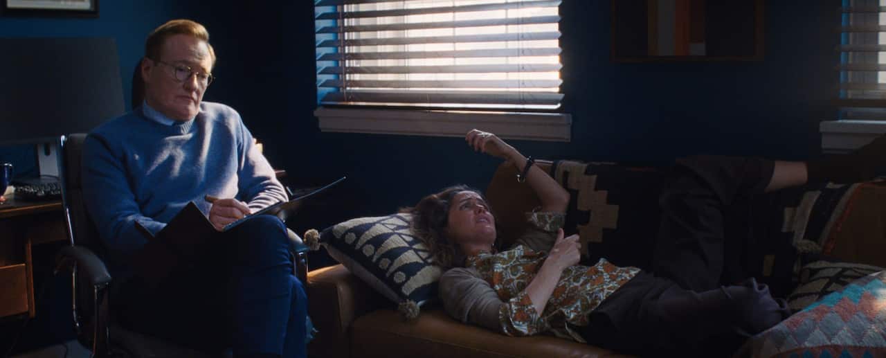 A woman lying down on a couch. A man is sitting next to her with a notebook.