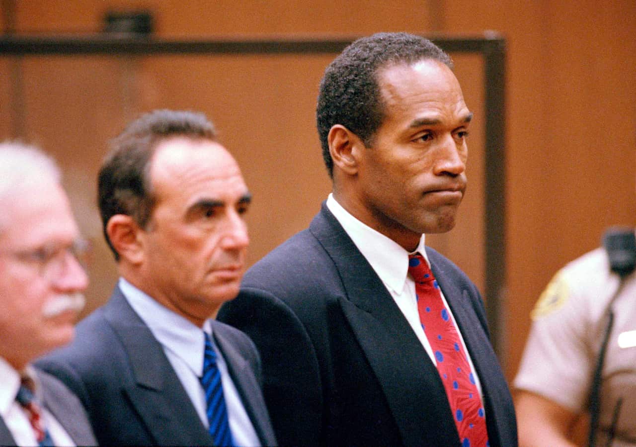 OJ Simpson standing in a court next to two lawyers in suits.