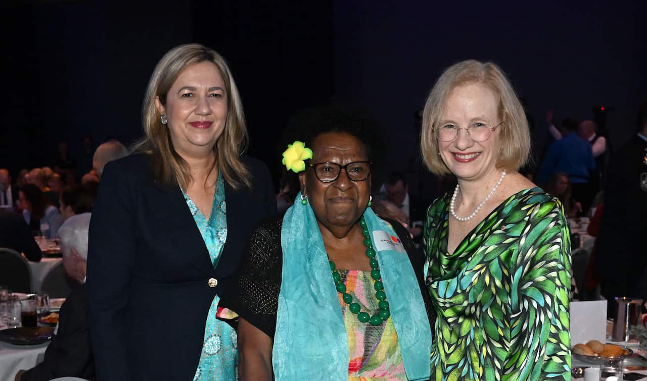 Two white women flank a third Torres Strait Islander woman, who wears a cyan scarf and a yellow frangipani behind her ear. The three smile at the camera in a function room with other guests in the background. 