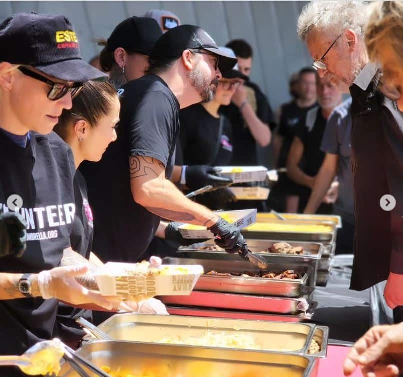 A row of silver dishes on tables with people serving on one side and people getting food on the other side.