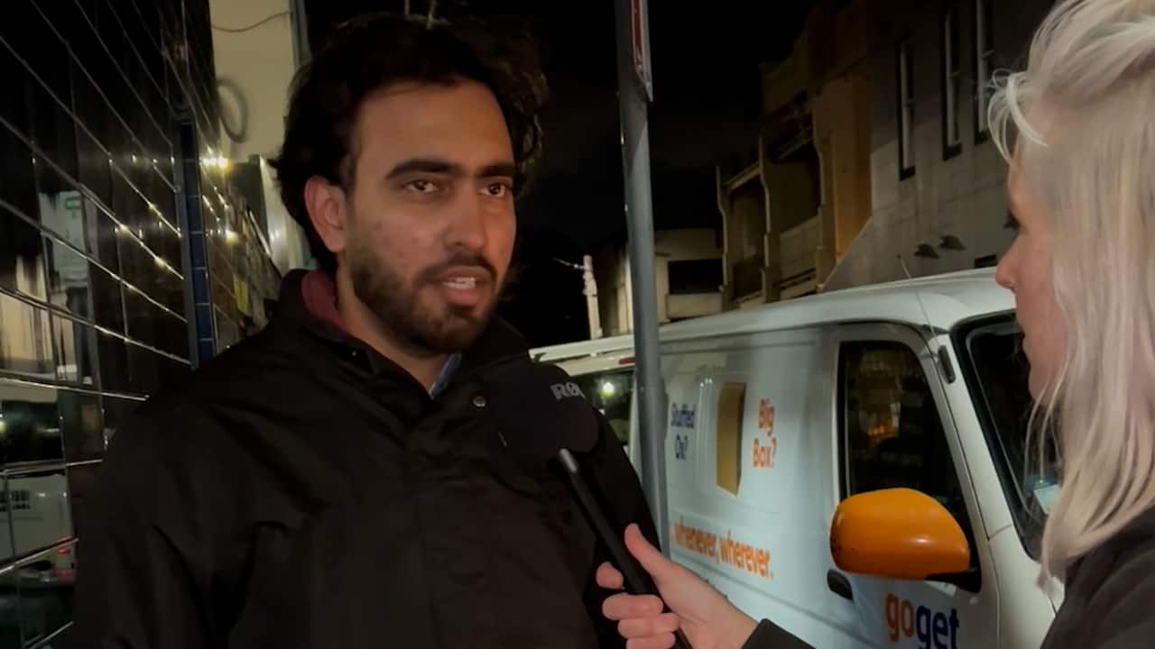 South Asian male delivery rider looks off to the distance while discussing delivery riders rights with caucasian woman. 