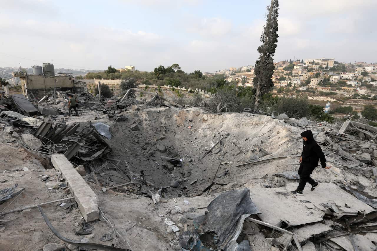 A wide shot shows a massive crater surrounded by rubble and debris in a residential area, with a person in dark clothing walking near the edge and houses visible on a distant hillside.