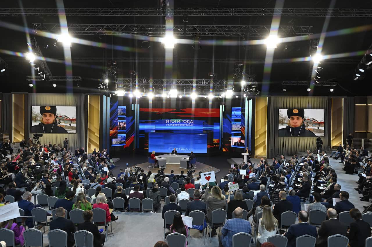 People seated inside a large room watching someone seated at a desk on stage speak.