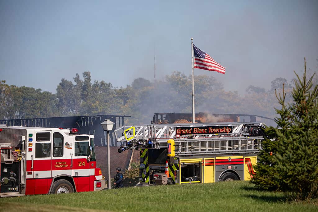 Fire engines group outside a burnt-out church with a sign that says Grand Blanc Township and an American flag flying.