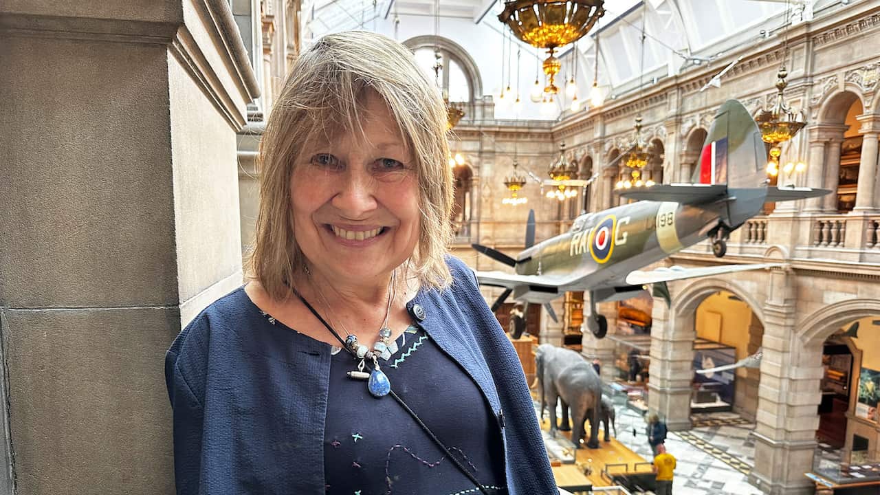 A woman with blonde hair posing for a photo in a museum. There is a World War Two fighter plane suspended from the roof behind her 