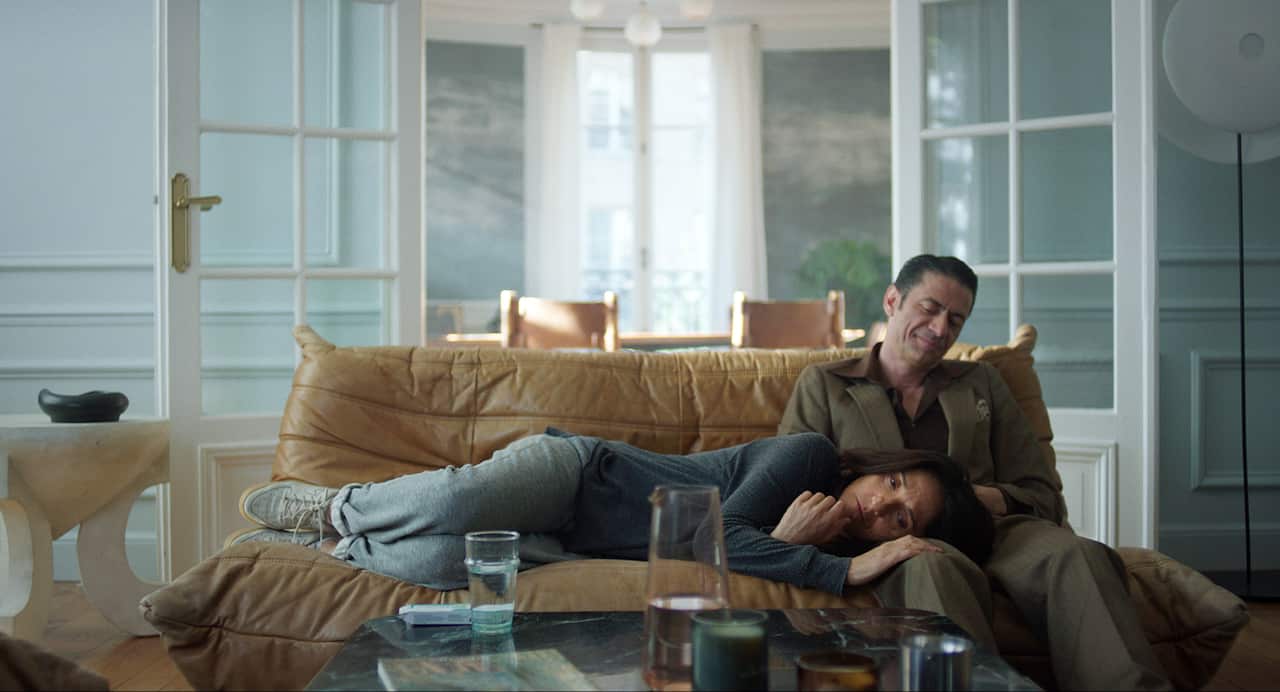A man in a brown suit sits on a couch. A woman lies on the couch, with her head in his lap.