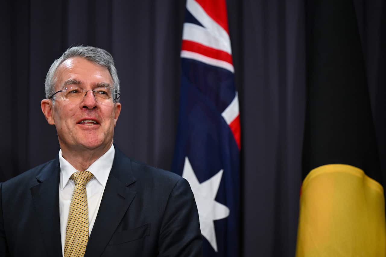 Man in suit speaks in front of Australian and Aboriginal flag.