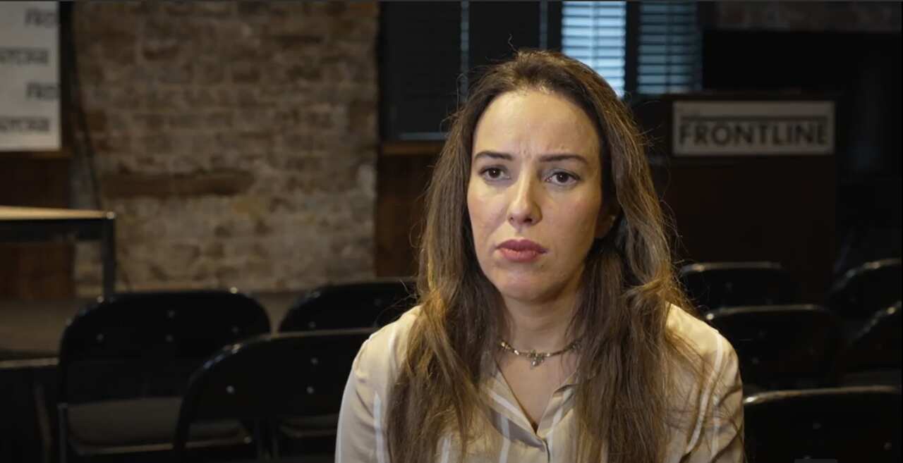 A woman is seated in front of a camera speaking to a journalist.