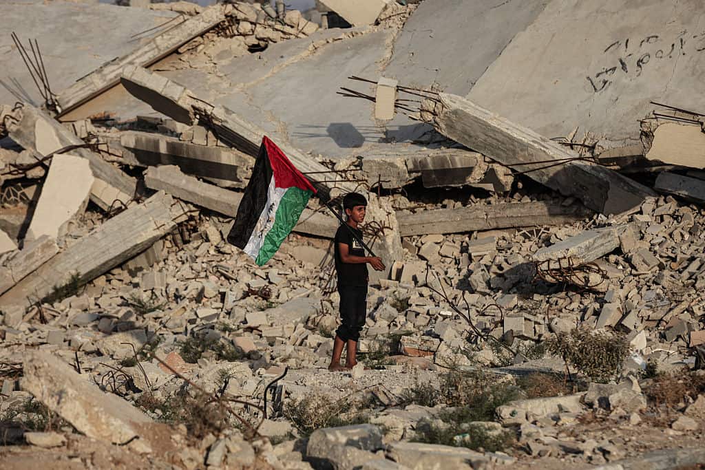 A boy stands amid rubble holding a Palestinian flag.