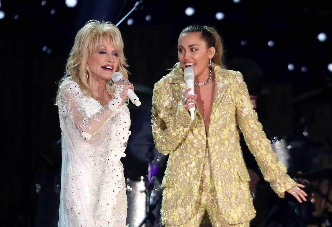 Two women - Dolly Parton and Miley Cyrus - wearing a white dress and old suit respectively, sing together.