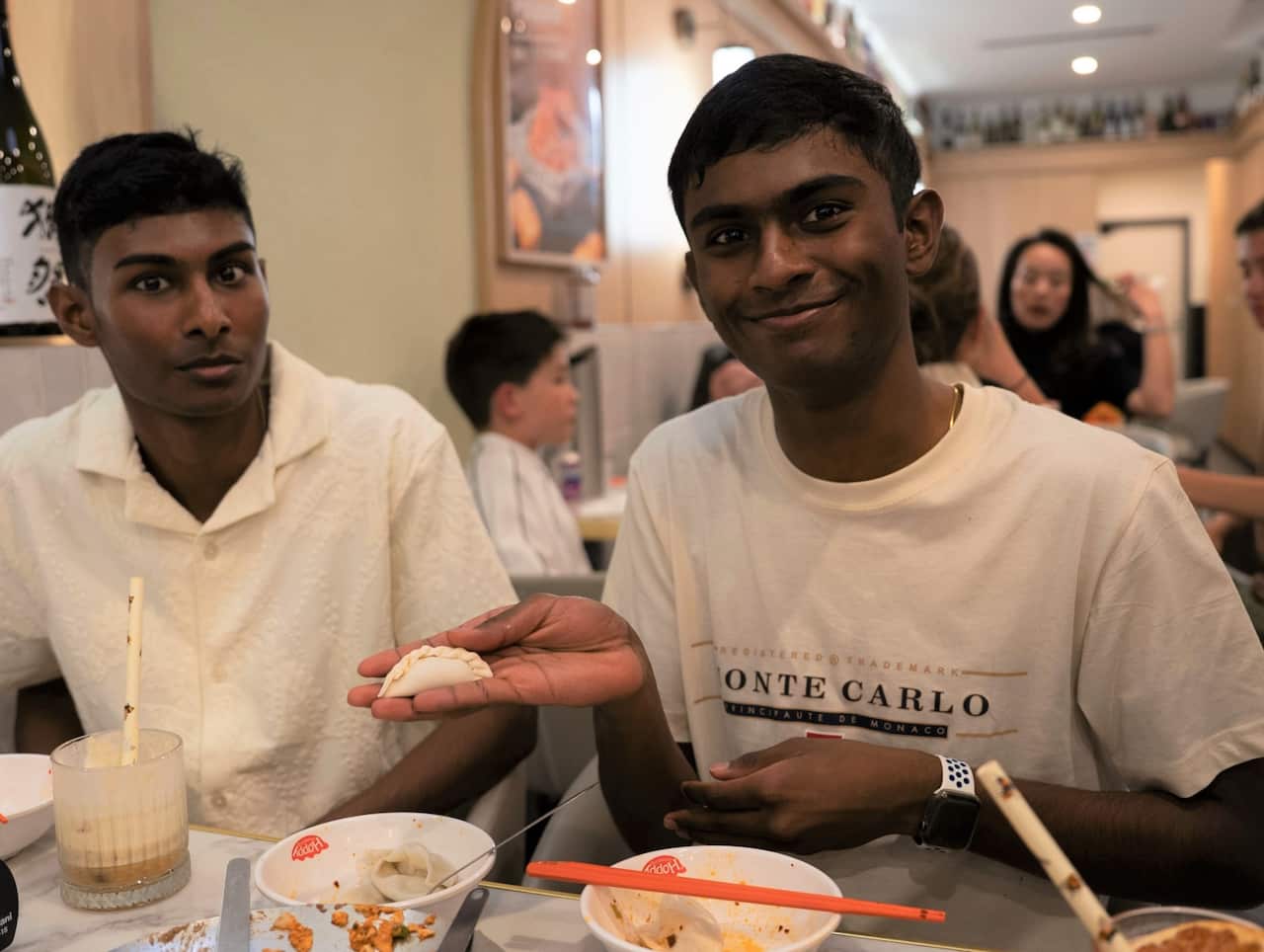 Two men in white shirts sit a a restaurant table, one holding a freshly made dumpling. 