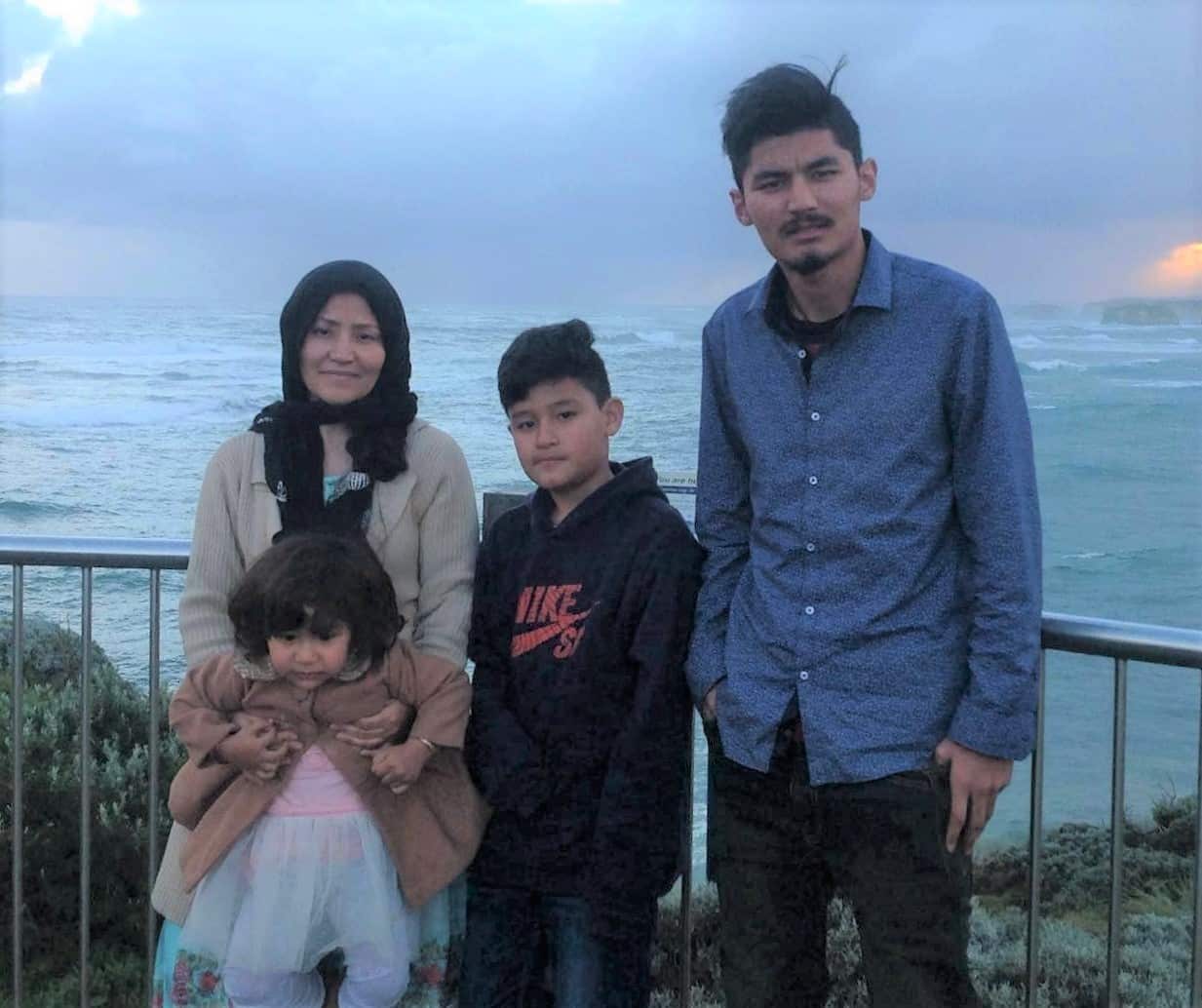 A man wearing a blue long-sleeved shirt stands at a fenced lookout with a woman and two children. The ocean is in the background. 