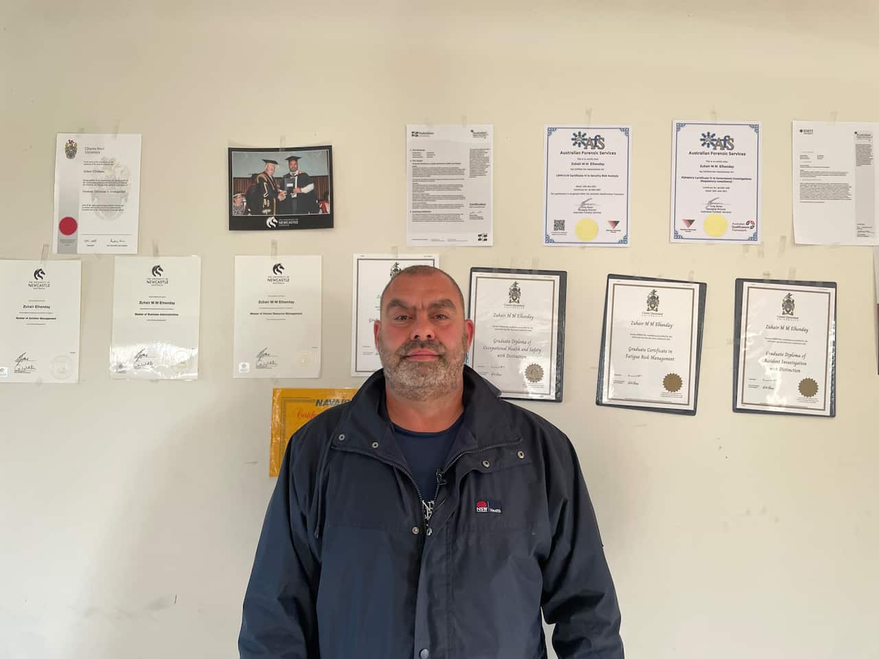 A man stands in front of university certificates stuck on a wall. 