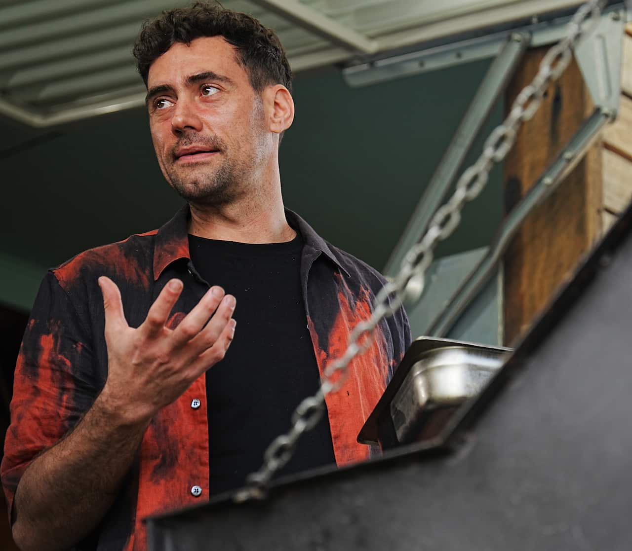 A man in a blue and orange shirt stands at a barbecue, gesturing with his hand.