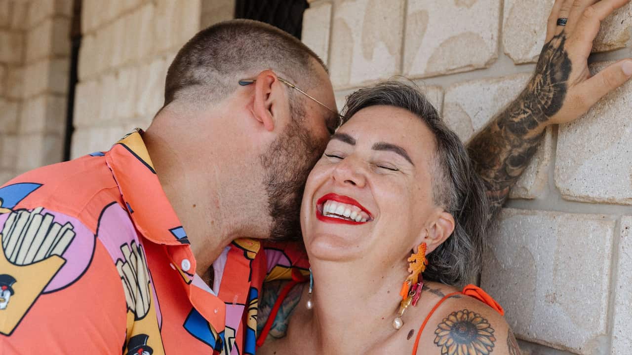 a man in a colourful party shirt leans against a wall and kisses the cheek of a smiling woman in red lipstick