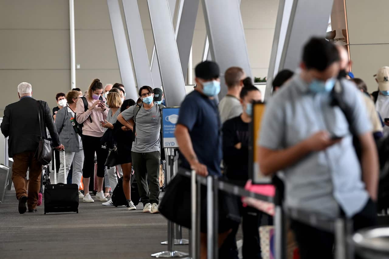 People wait in a queue at an airport terminal, with many people wearing light blue masks on their face.