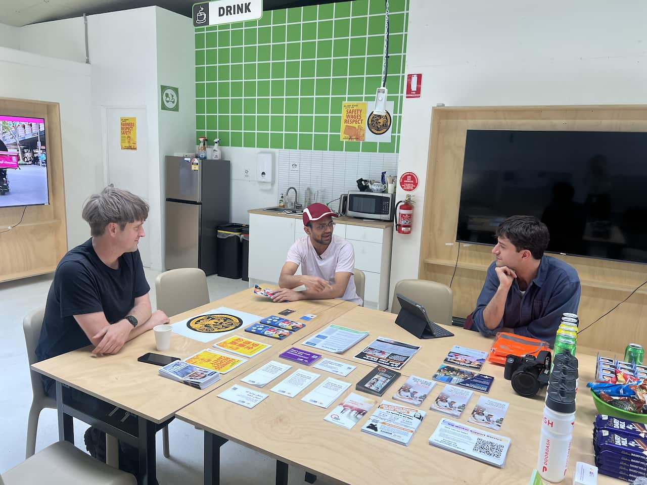 Three men sit around a wooden table with pamphlets sprawled in front of them.