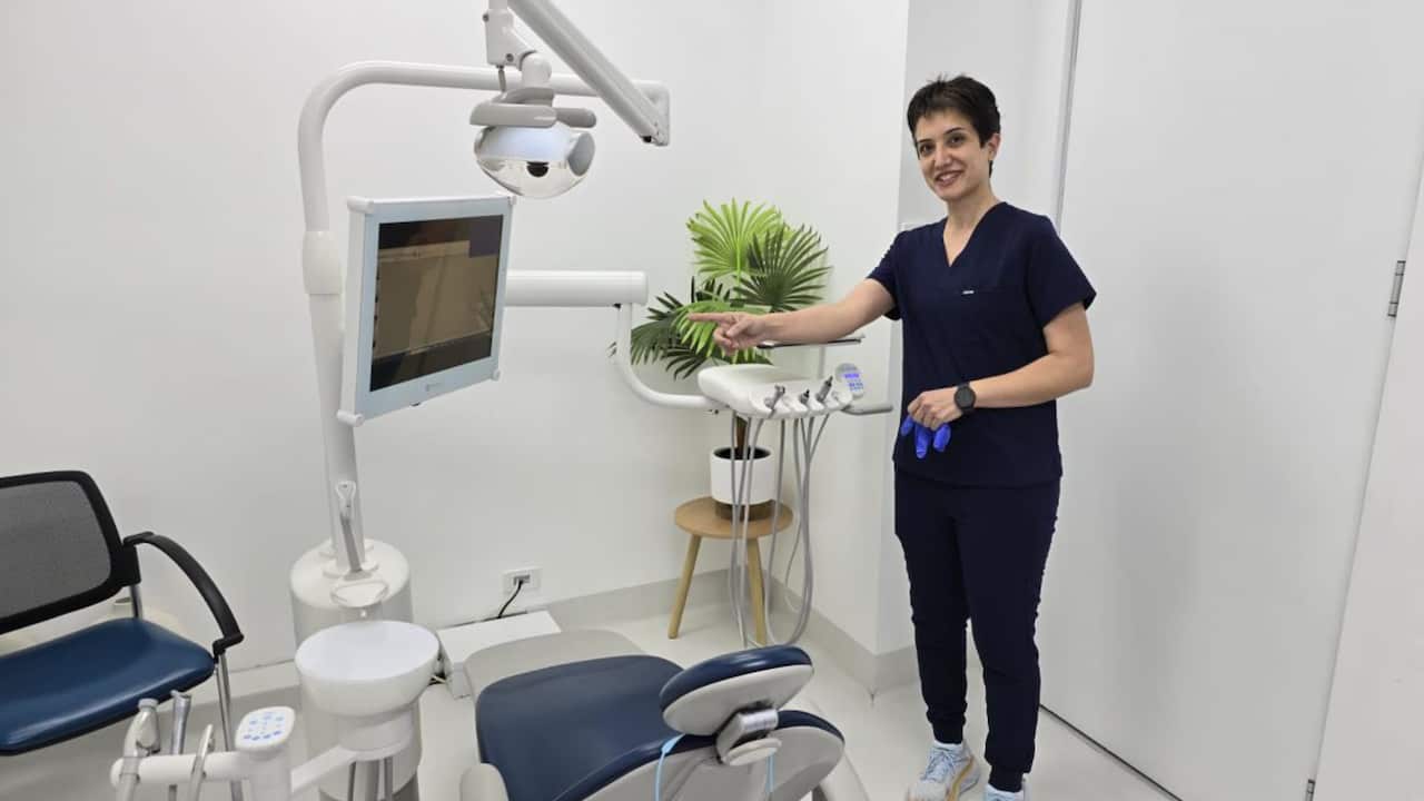 A woman in navy scrubs standing in a dentist's room, smiling and pointing toward a monitor beside the patient chair.