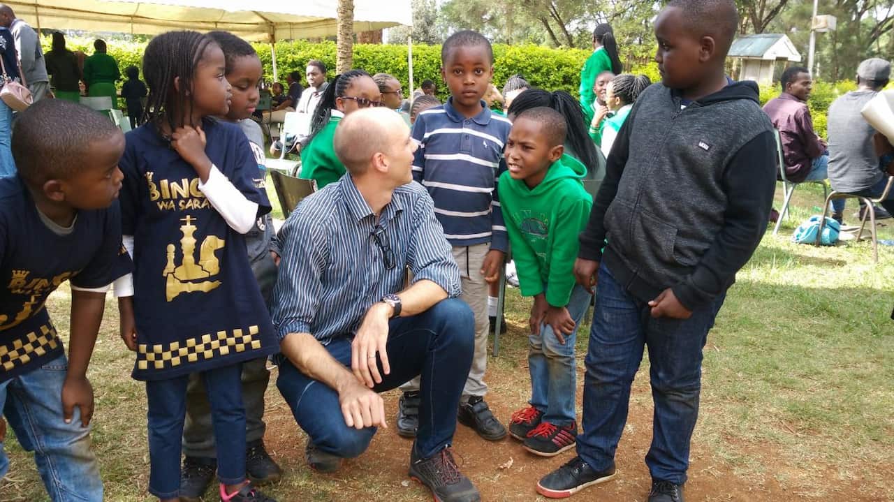 A man in a striped shirt and blue jeans is crouching in the garden next to a group of young children who are standing.